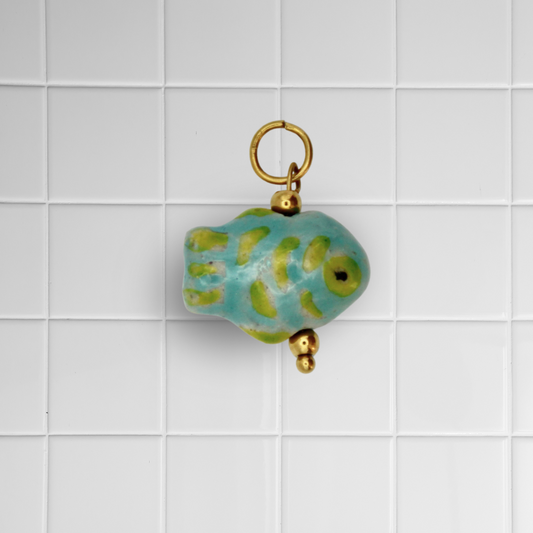 Grün-gelber Keramikfisch-Anhänger mit goldener Öse auf weißem Kachel-Hintergrund