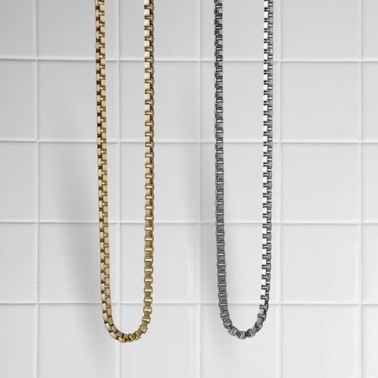 Zwei Box Chain Ketten in Gold und Silber vor weißem Kachel-Hintergrund