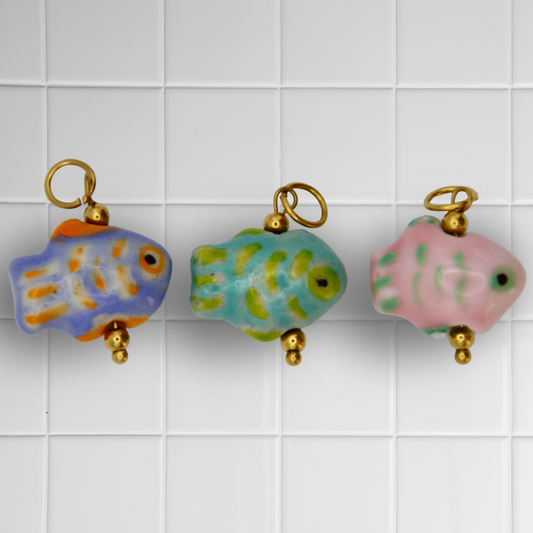 Drei Keramikfisch-Anhänger in Blau, Grün und Rosa mit goldenen Details auf weißem Kachel-Hintergrund