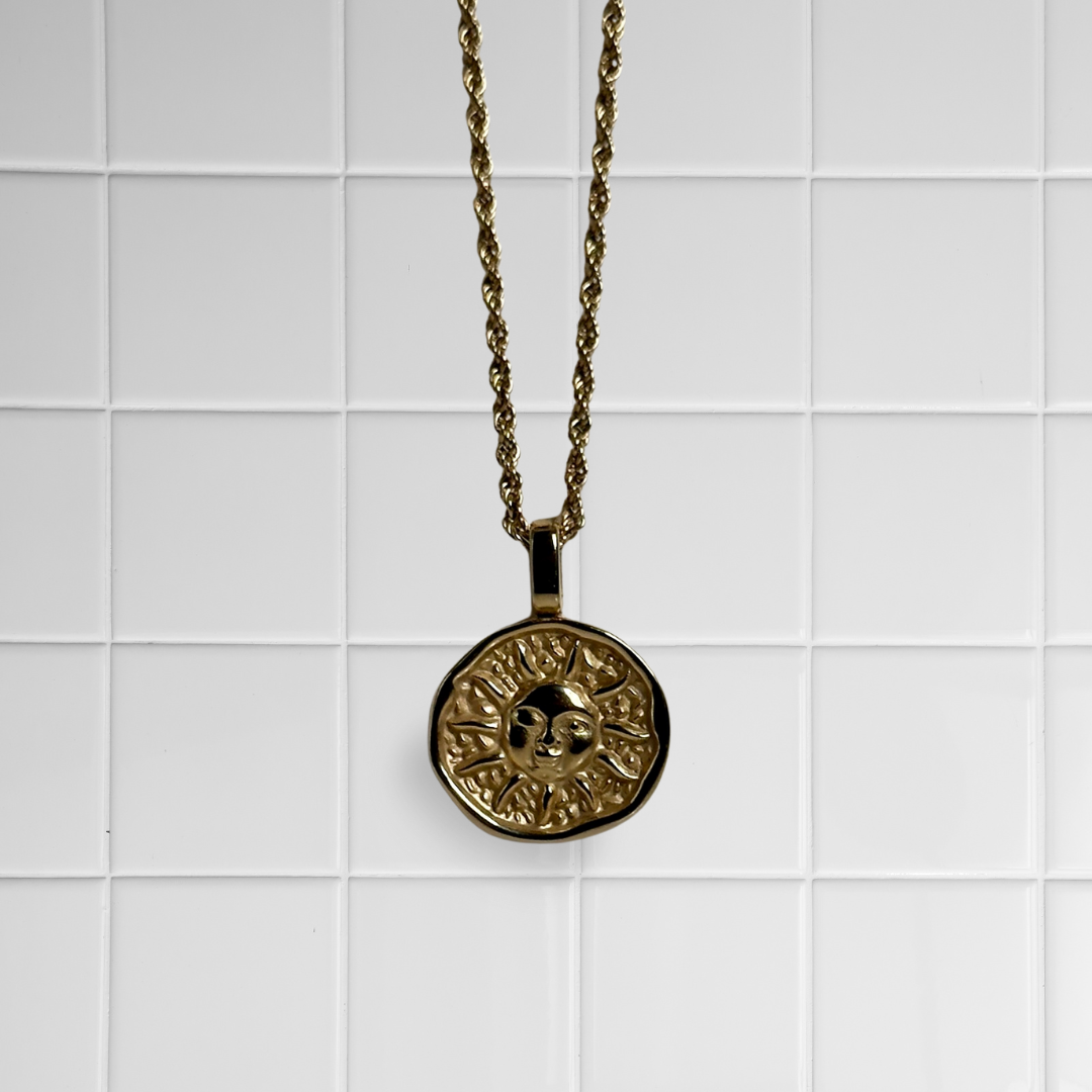 Goldener Sonnen-Anhänger an gedrehter Kette vor weißem Kachel-Hintergrund