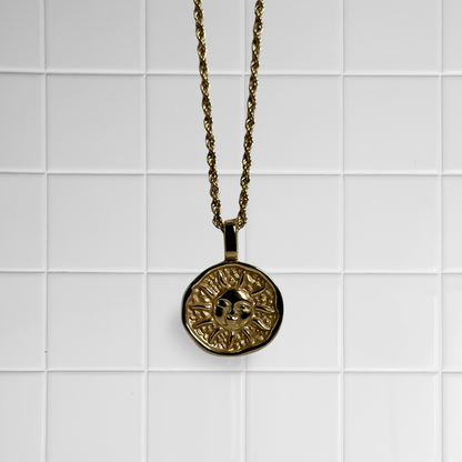 Goldener Sonnen-Anhänger an gedrehter Kette vor weißem Kachel-Hintergrund